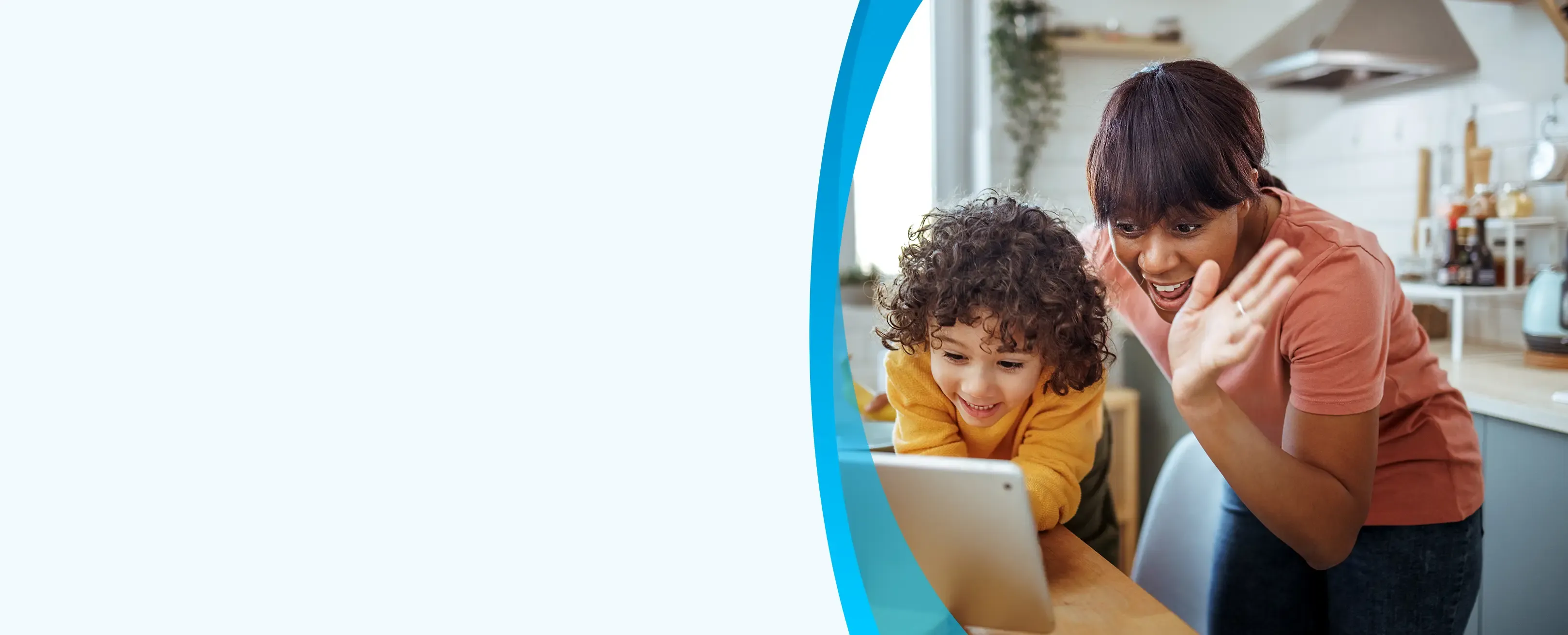 A smiling mother waves at a tablet screen while a young child watches during a video call in the kitchen.